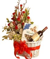 A basket of red wine, a box of chocolate, and bouquet of red & white carnations